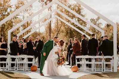 A bride and groom share a kiss under an outdoor wedding archway, surrounded by guests.