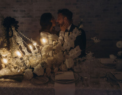 A newlywed couple kissing while sitting at a reception table.