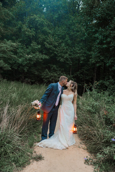 A bride and groom stand on a dirt path sharing a kiss. The bride is in a white strapless wedding down and the groom is in a nave blue suit with a pink tie. It is dark out and they are carrying lanterns to light the way. They are surrounded by tall grass and lush green woods.