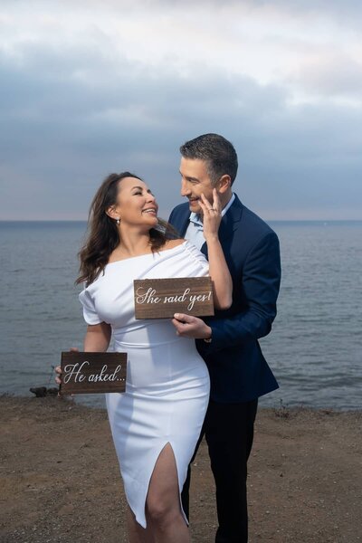 Engaged couple celebrates with “He asked, she said yes” signs during coastal engagement proposal session at Roessler Point in Palos Verdes