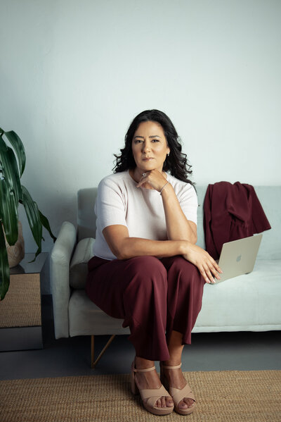 Asmahan sitting confidently on a modern sofa with a laptop in her lap, styled in burgundy trousers and nude heels, exuding calm authority in a minimalist workspace