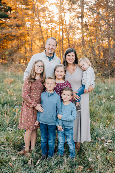 Family smiling together near vibrant fall trees, wearing cozy autumn outfits.