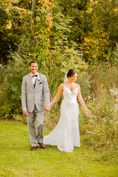 Bride and groom walking holding hands while bride touches wild flowers