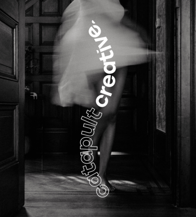 Black and white artistic image featuring a blurred figure of a woman in motion wearing a flowing dress, standing in front of a wooden paneled door. The bold, curved text 'Catapult Creative' is displayed vertically along the right side of the image, blending modern typography with the classic atmosphere of the room.