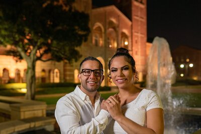 Engaged couple holding hands with joyful expressions at night in front of the iconic UCLA building in Los Angeles, showcasing fine art portrait photography