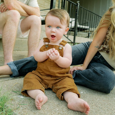 Close up image of a baby sitting on the ground at his parents' feet wearing overalls