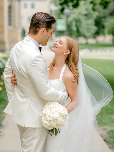 Bride with a cathedral veil holds groom in a white tux around the waist with church facade behind them as they look into each other's eyes.