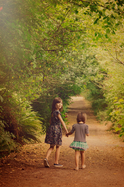 two sisters walking on a trail in the woods holding hands, the oldest sister is looking back at the camera
