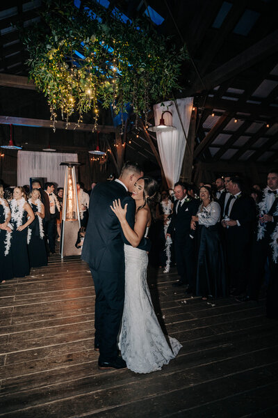 woman in black tux hugging and dancing with a woman in a white dress on wooden floor with vines and lights hanging from the ceiling