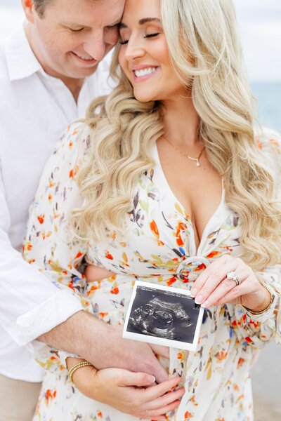 Couple hugging at Lighthouse Beach holding up sonogram picture. 