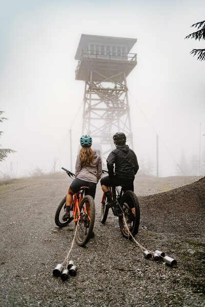 an eloping couple on mountain bikes looking up towards a fire lookout,