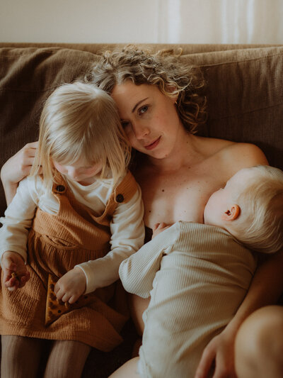 en mamma ligger hemma i sin soffa och håller om sin dotter och med sin son som ammar