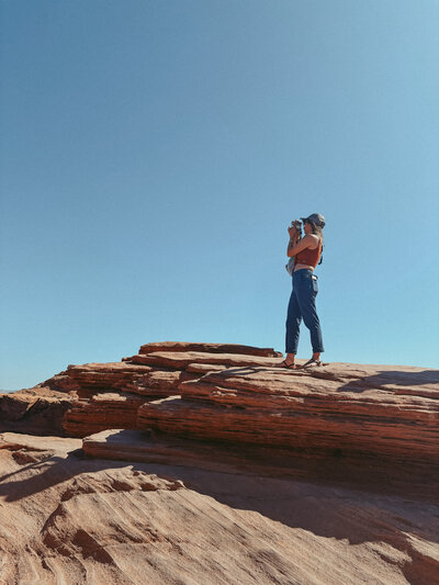 Elopement photographer standing on red rock formations in Page, Arizona, overlooking Horseshoe Bend. She holds a film camera under a clear blue sky, capturing the desert landscape in a candid, adventurous moment.
