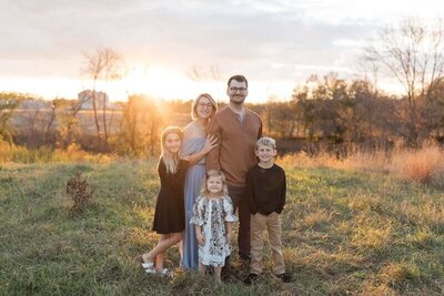 A family of five stands together in a grassy field at sunset, smiling warmly with golden light in the background.