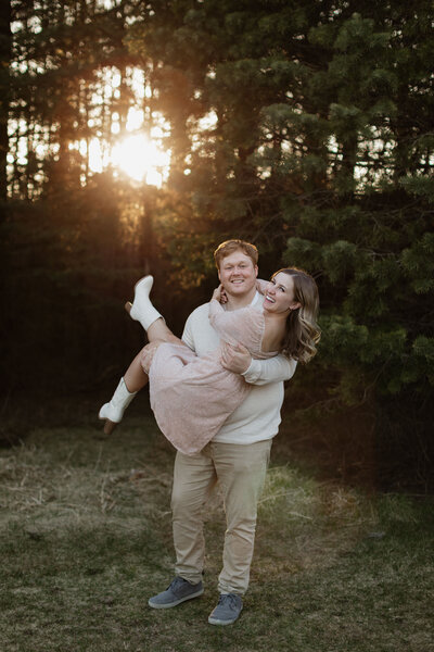 man holding woman in his arms while they both smile and gentle sunlight shines through the trees