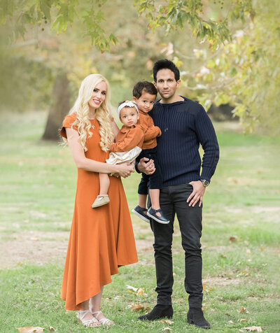 Mom, baby and son wearing burnt orange standing smiling with dad in the park by Los Angeles newborn photographer