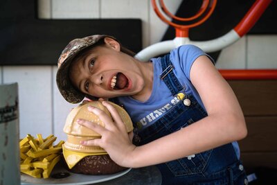 An energetic young child in denim overalls and a camo hat playfully pretends to bite into a giant burger, with eyes wide in excitement, during a fun-filled family photo shoot in Connecticut.
