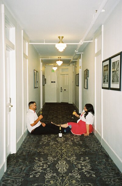 Couple sitting in a hotel hallway drinking champagne during their downtown engagement photos captured on film