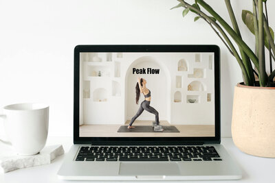 Sarah demonstrates a yoga pose for a 'Peak Flow' class on a laptop screen, placed on a white desk next to a cup and a potted plant, in a room with a white, minimalist interior.