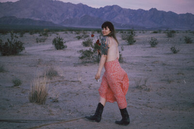 A person wearing a red skirt, boots, and a floral top stands in a desert landscape with mountains in the background, evoking the creative flair of Sweet Pea Ink Creative’s brand design.