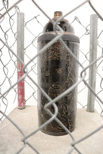 A dark ceramic spray paint can is decorated with a carving of young children and sits atop a graffiti painted pedastal, but is caged in by a wire fence