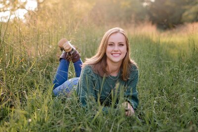high school senior girl in sweater jeans and boots laying on stomach in tall green grass