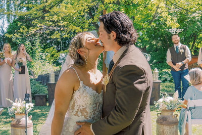 Bride and groom sharing their first kiss under the trees during a romantic outdoor wedding ceremony in Idaho, surrounded by friends and floral details.
