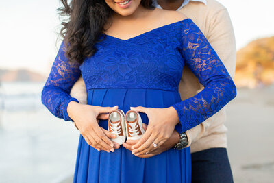 Pregnant woman holds baby shoes in front of her stomach while her partner embraces here from behind.