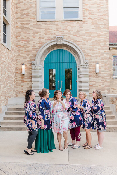 Bride and Bridesmaids wearing dark blue robes with bright pink and green leaf floral pattern, giggling while standing outside of St. Louis Wedding Venue during Bridal Portraits | Evalyn & Co. Photography