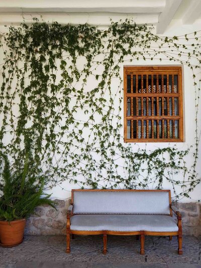 Wooden bench with gray upholstery beneath climbing vines and a traditional wooden window on a white wall in Cusco, Peru ©Stephanie Dosch | theViatrix Luxury Travel