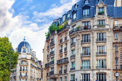 Elegant historic apartment buildings with ornate facades and mansard roofs line a Parisian street. Balconies adorned with greenery are visible under a clear blue sky with scattered clouds. A tree is partially visible on the left.