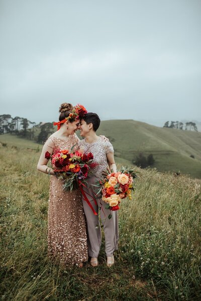 Two brides embracing on the top of a hill in South Gippsland holding colourful bouquets
