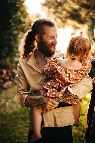 Tattooed father holds his toddler daughter in a field at sunset.