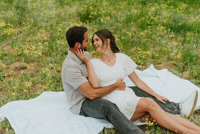 A man is holding the waist of his fiance as she sits between his legs. She's holding the side of his face with one hand and their smiling at each other. Their sitting on a white blanket in a field of small yellow flowers.