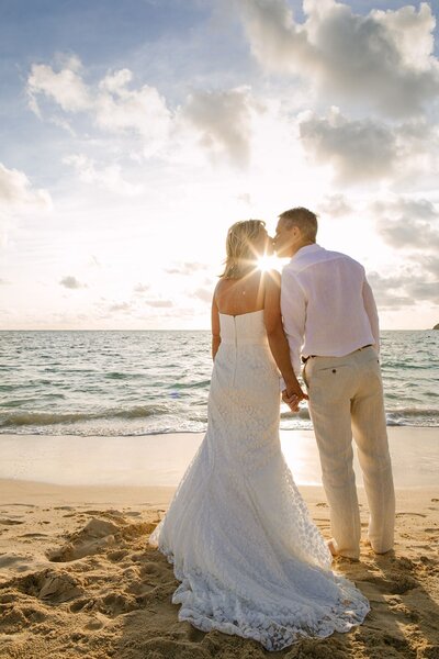 A man and woman hold hands kissing on the beach in Hawaii.