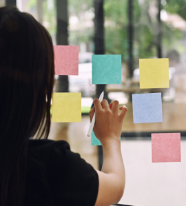 Woman placing colorful sticky notes on a glass wall, illustrating employee engagement, idea-sharing, and team planning.