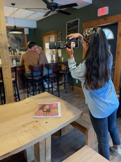 A person with long dark hair, embodying the New England lifestyle photographer vibe, captures a couple sitting closely in a cozy, rustic bar. A newspaper rests on a wooden table in the foreground, while an exit sign and ceiling fan hint at the room's characterful charm.