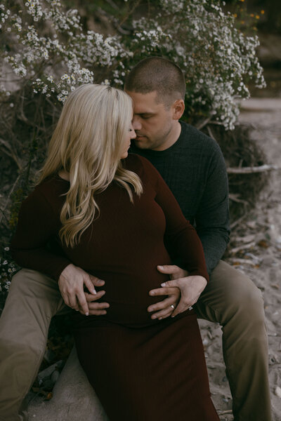 Maternity photoshoot, dark and moody outdoors in front of white wildflowers with mom sitting on dad's lap turning toward him and their heads are together