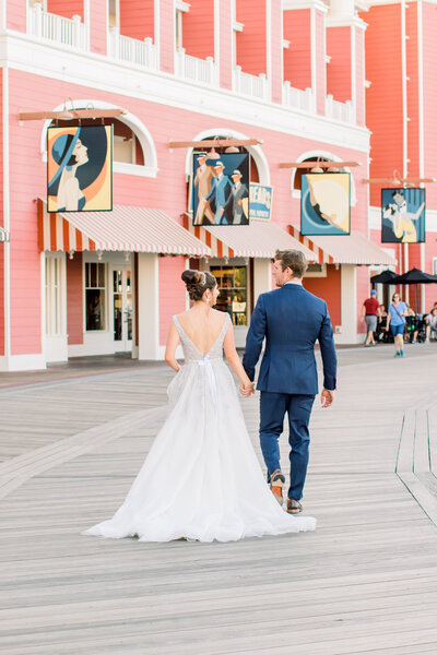 Bride and groom holding hands walking along the colorful Disney Boardwalk in wedding attire.
