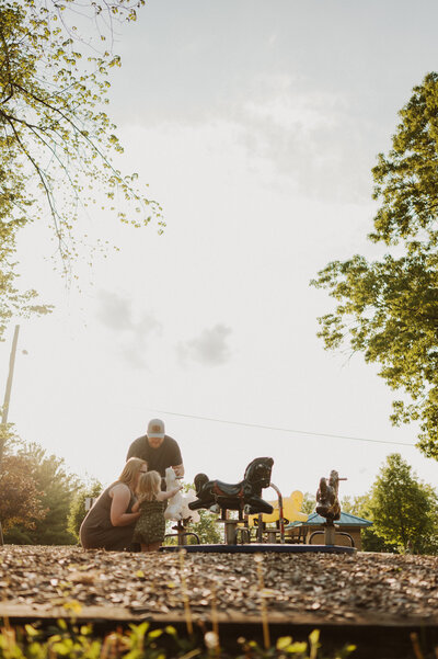 A family with a child at a playground with a merry-go-round in a park.