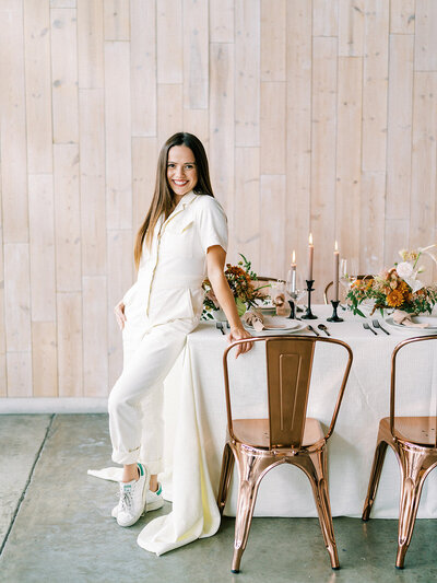 Woman in a white jumpsuit posing next to a table set with florals and candles and copper chairs