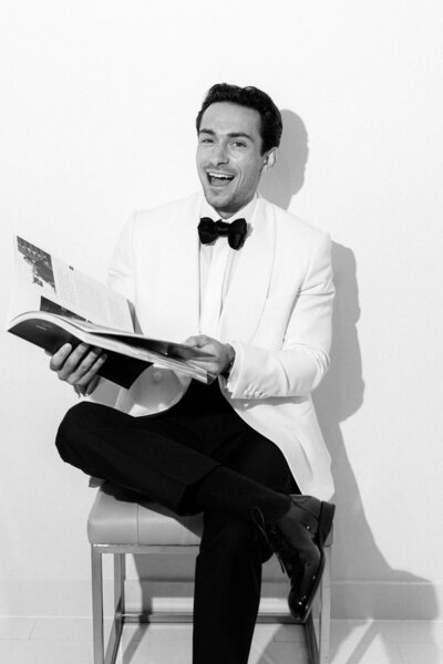 Black and white image of a smiling groom wearing a white tux holding an open magazine