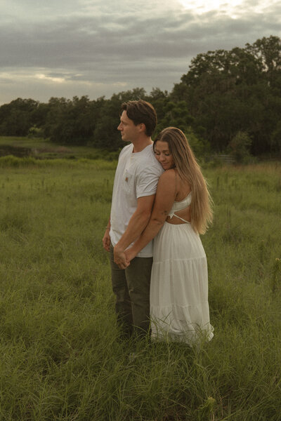 Woman resting her head on her partner’s shoulder as they hold hands and stand in a peaceful grassy field.