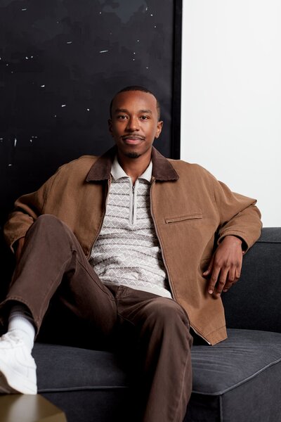 Dennis, a young black man with short hair, sitting on a grey coach. He is wearing a brown suede jacket over a white polo shirt and brown jeans.