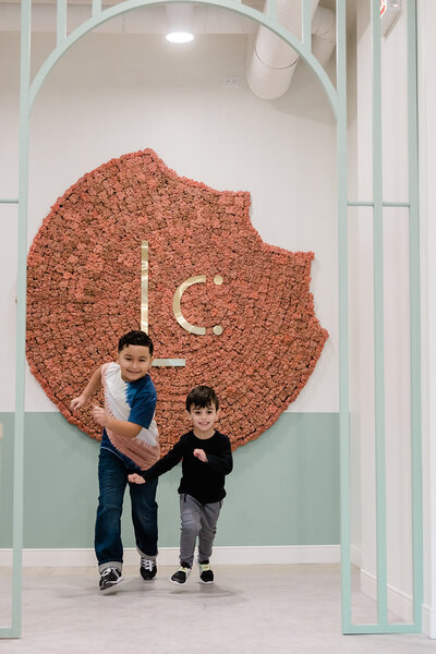 Two casually dressed boys, with dark hair, are captured in motion, looking like they're about to run right for you. The boys are smiling and are framed by the soft, minty green architectural details of Chicago pediatric dentistry office Little Chompers. In the background is  a beautiful, artistic wall fixture featuring Little Chompers eye-catching logo.