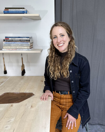 A smiling artist in a dark blue work coat and corduroy pants sits in a printmaking studio with art books and printmaking tools in the background.