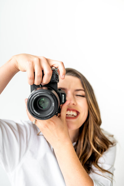 A close up shot of Kala from the chest up. She is holding her camera to her face with it pointing at the camera and smiling.