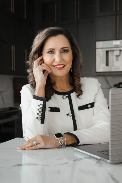 A woman with long brown hair, wearing a white and black blazer, sits at a marble table with a laptop, lightly touching her face and smiling.