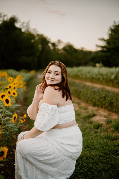 Smiling senior girl crouched next to sunflowers wearing white dress in Annapolis Maryland.