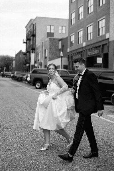 Bride lifting her dress as she walks across the street with her groom during downtown Holland wedding portraits.
